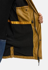 Giacca impermeabile marrone con un esterno liscio, fodera in fleece nera, dettagli gialli e numerose tasche all'interno. Chiusura con cerniera.