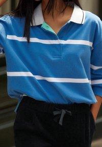 Niebieska koszulka polo w białe poziome paski, z jasnoniebieskim kołnierzykiem i plisą na guziki, w komplecie z ciemnymi spodniami. Miękki, casualowy materiał.