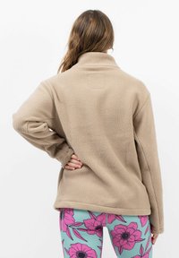 Beiger Fleece-Pullover mit hohem Kragen und strukturierter Oberfläche, kombiniert mit lebhaften Leggings im Blumenmuster in Blau und Pink.