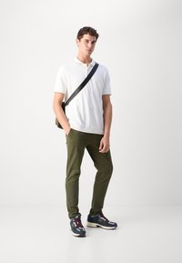 Hvid polo shirt, olivengrøn slim-fit bukser og multicolor New Balance sneakers. Model bærer en sort skuldertaske. Simpelt design.