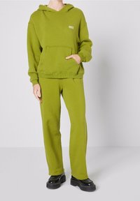 Felpa verde con tasca frontale e polsini a costine, abbinata a pantaloni verdi coordinati. L'outfit presenta un tessuto testurizzato e una vestibilità comoda.