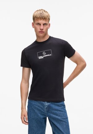 T-shirt en coton noir arborant un logo rectangulaire blanc avec "KARL LAGERFELD JEANS." À manches courtes et col rond, associé à un jean bleu.