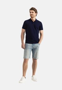 Marineblauw poloshirt met korte mouwen, gecombineerd met lichtblauwe denimshorts. Het model draagt witte sneakers en staat voor een effen achtergrond.