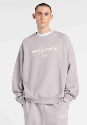 Junger Mann trägt ein helllila New Balance Athletics Sweatshirt und passende Jogginghose, steht mit den Händen in den Taschen.