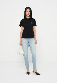 Μαύρη βαμβακερή μπλούζα με στρογγυλή λαιμόκοψη και λογότυπο μπροστά, συνδυασμένη με ανοιχτό μπλε τζιν, μαύρες μυτερές γόβες και άσπρη φουσκωτή τσάντα.