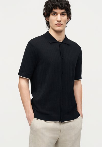 Νεαρός άνδρας με σγουρά μαλλιά, που φοράει μαύρο κοντομάνικο πουκάμισο με κουμπιά και ανοιχτό μπεζ παντελόνι, στέκεται με τα χέρια στις τσέπες.