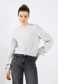 Felpa grigia chiara con scollo rotondo e maniche lunghe leggermente a sbuffo, abbinata a jeans neri a vita alta. Morbida al tatto, vestibilità casual.