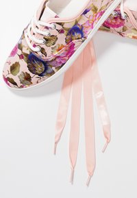 Baskets à motif floral rose avec des fleurs violettes et dorées, semelle blanche, et longs lacets en ruban satiné rose pâle sur fond blanc.