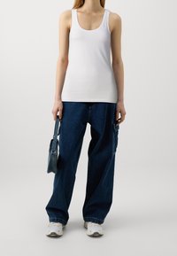 Mujer vestida con una camiseta sin mangas blanca, pantalones cargo de mezclilla oscuros y sueltos, zapatillas blancas, sosteniendo un pequeño bolso de mezclilla frente a un fondo liso.