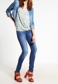 Veste en denim sur un top floral clair, associée à un jean skinny bleu et des sandales à talons hauts en suède marron. Look texturé et superposé.