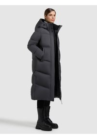 Dlouhý černý péřový kabát s kapucí, s prošívaným designem, dvěma bočními kapsami a plným zipem. V kombinaci s černými šněrovacími botami.