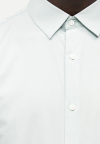 Κοντινό πλάνο καφέ επιδερμίδας που φοράει ένα τραγανό λευκό πουκάμισο με γιακά και τρία ορατά λευκά κουμπιά, κουμπωμένο μέχρι τον λαιμό.