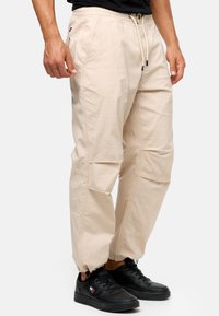 Beige Cargohosen mit elastischen Bündchen, Front- und Seitentaschen, verstellbarem Kordelzug an der Taille, aus leichtem Stoff gefertigt. Schwarze Sneakers getragen.