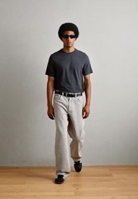 T-shirt gris à manches courtes, jean gris clair, ceinture noire et baskets noires. Le modèle porte des lunettes de soleil ; cheveux coiffés en afro.
