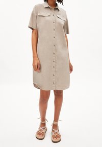 Robe chemise beige à manches courtes avec patte de boutonnage, deux poches poitrine, ourlet arrondi et texture douce. Associée à des sandales beiges à brides.