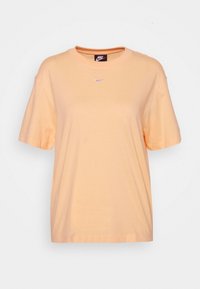 Světlá oranžová tričko s krátkým rukávem Nike, s malým fialovým logem swoosh umístěným uprostřed pod kulatým výstřihem na jednobarevném pozadí.