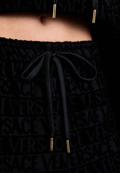 Haut court noir en tissu texturé arborant le mot "VERSACE" dans un motif répétitif, terminé par des extrémités de cordon de serrage accentuées en or.