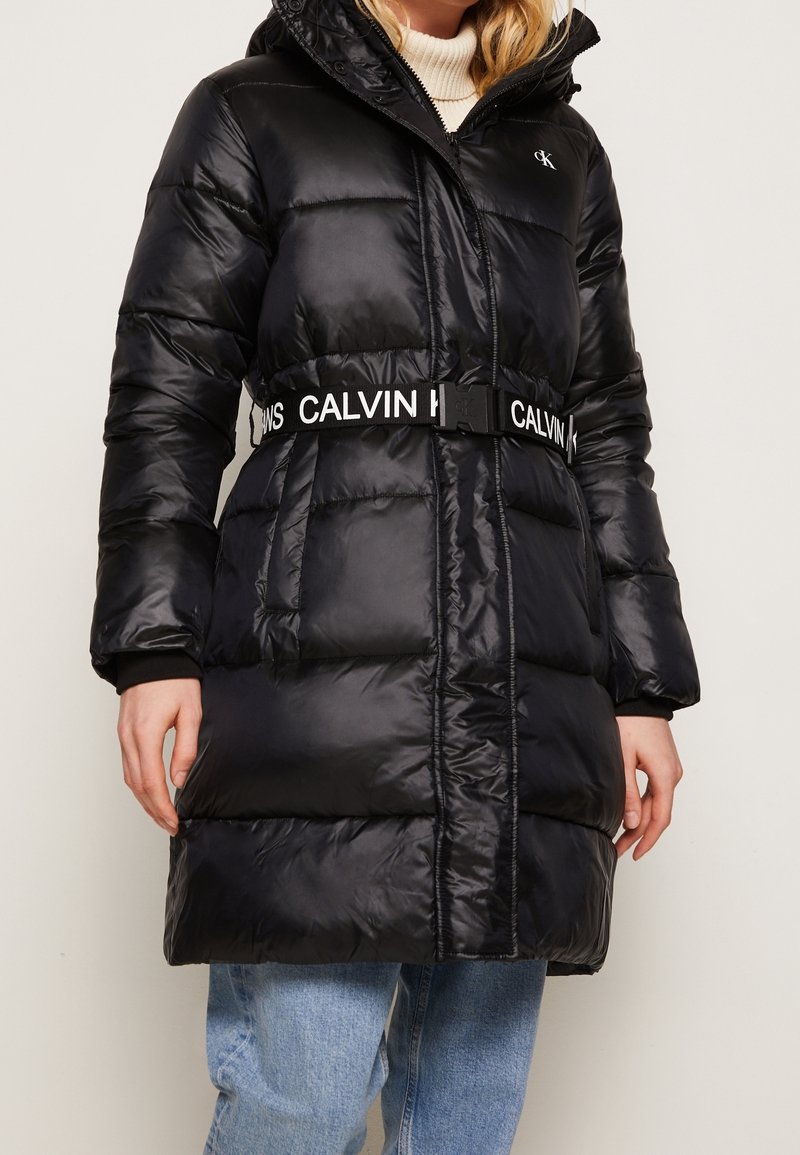 Schwarzer Steppmantel mit Kapuze, Taillengürtel mit dem Schriftzug "CALVIN", glänzendem Finish und gerippten Bündchen. Midilanger Schnitt.