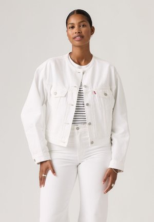 Donna con capelli scuri indossa una giacca e pantaloni di denim bianchi sopra una maglia a righe bianche e nere, in piedi davanti a uno sfondo chiaro e uniforme.