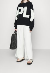 Sudadera negra con grandes letras blancas "RPLY", combinada con pantalones blancos holgados, un bolso negro texturizado y sandalias blancas gruesas con detalles florales.