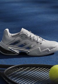 Biela tenisová obuv Adidas so striebornými pruhmi na modrej kurte vedľa čiernej tenisovej rakety a žltej tenisovej loptičky.