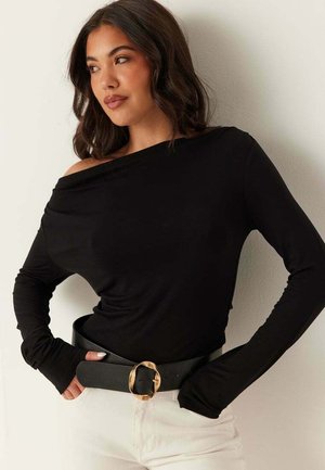 Kvinde med skulderlangt mørkt hår iført en sort off-shoulder langærmet top, bredt sort bælte med guldlås og hvide bukser.