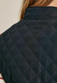 Funkcionális, sötétkék, steppelt mellény gyémántmintával, magas gallérral, és a vállnál hímezett "Joules" felirattal. Puha, texturált anyag.