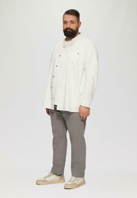Man met een baard, gekleed in een wit overhemd, grijze broek en beige sneakers, staande tegen een effen lichte achtergrond.