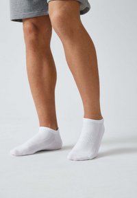 Weiße Knöchelsocken mit strukturiertem Muster, weichem Stoff und elastischen Bündchen, die auf einem Bein über einem grauen Kleidungsstück platziert sind.