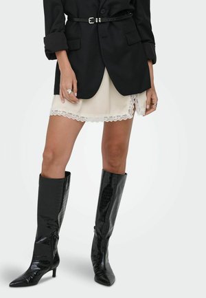 Kobieta w czarnym blezerze z paskiem na kremowej mini spódnicy z koronkową lamówką i czarnych, sięgających kolan, spiczastych butach.