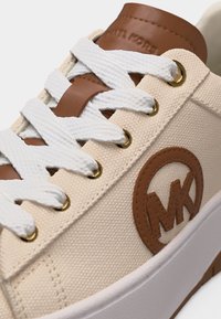 Μπεζ πάνινο αθλητικό παπούτσι με λευκά κορδόνια, καφέ δερμάτινη γλώσσα και λογότυπο, χρυσά οπές και λευκή ελαστική σόλα.