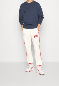 Person klädd i en marinblå Nike-tröja, vita Nike-sweatpants med röda ränder och vita sneakers med röda Nike-logotyper. Händerna i fickorna.