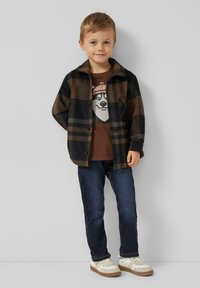 Koszula w kratę w odcieniach brązu i czerni, noszona na ciemnobrązowej koszulce z nadrukiem niedźwiedzia. Do tego niebieskie dżinsy i białe trampki.