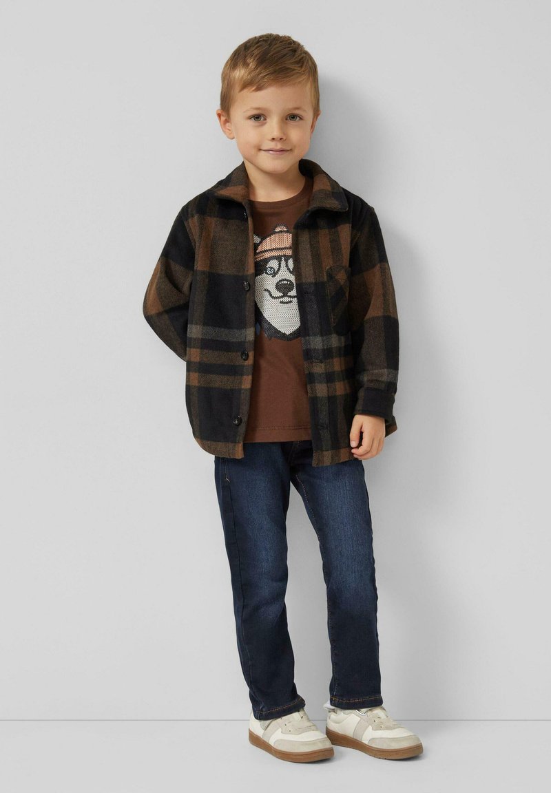 Koszula w kratę w odcieniach brązu i czerni, noszona na ciemnobrązowej koszulce z nadrukiem niedźwiedzia. Do tego niebieskie dżinsy i białe trampki.