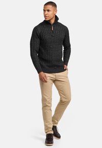 Dunkelgrauer, strukturierter Kabelstrickpullover mit einem Reißverschlusskragen, kombiniert mit beigen Hosen und braunen Schnürstiefeln.