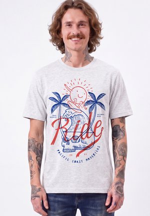 Homme avec des tatouages sourit, portant un t-shirt gris clair avec un crâne coloré, une vague, des palmiers, le soleil et un graphisme texte "Ride".