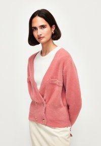 Cardigan rosa morbido con scollatura a V profonda, due tasche posteriori e dettagli con bottoni dorati, indossato sopra un maglione bianco. Tessuto liscio.