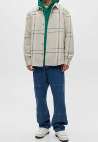 Beige geblokt overhemdjack met bruine knopen over een groene hoodie. Losse blauwe denim jeans met horizontale stiksel. Witte sneakers.