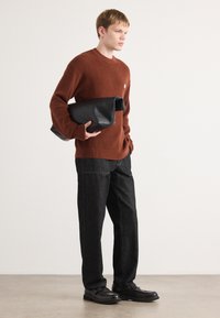 Roestkleurige gebreide trui, donker denim broek met stikdetails, en zwarte leren clutch. Zwarte schoenen met textuurelementen.