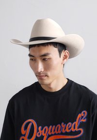 Chapéu de cowboy de feltro bege com aba larga e uma faixa preta, usado com um moletom preto com letras coloridas. Textura suave e design estruturado.