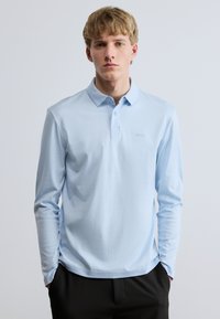 Φωτεινό γαλάζιο μπλουζάκι πόλο με μακριά μανίκια και κλειστό γιακά, με διακριτική κεντημένη λογότυπο στο αριστερό στήθος. Λείο ύφασμα και εφαρμοστό σχέδιο.