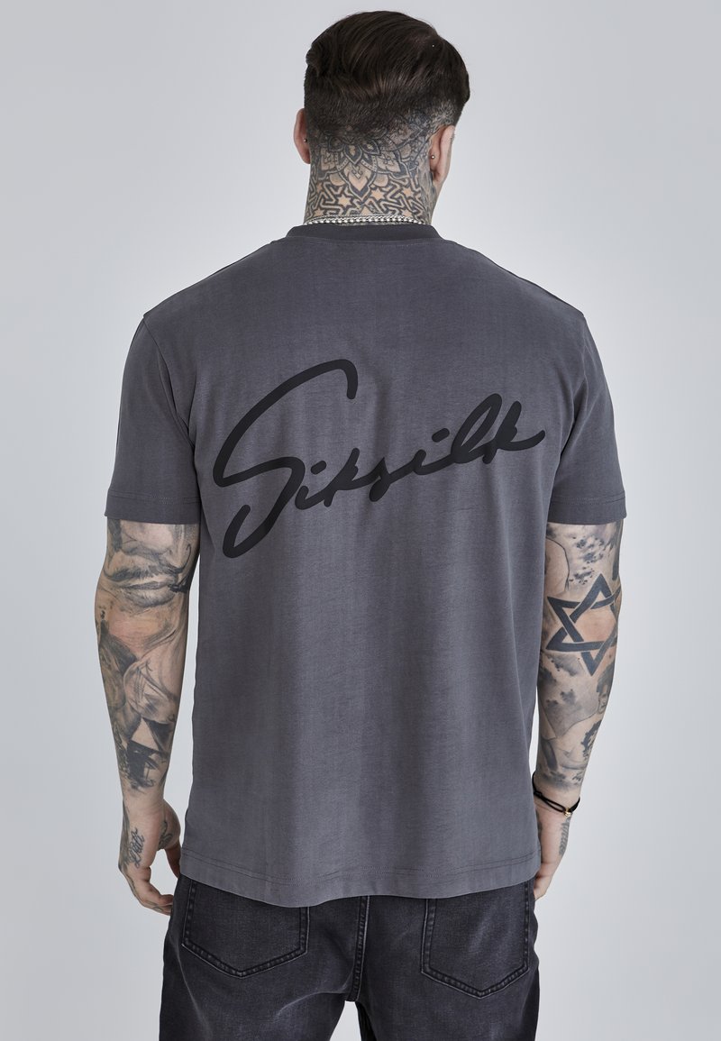 Grijze katoenen t-shirt met een zwart cursief "Sisilk" logo op de achterkant. Korte mouwen en een klassieke snit. Draagt met donkere jeans.