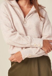 Pullover rosa chiaro, testurizzato, con colletto a mezzi zip e polsini a costine, arricchito da bottoni dorati. Indossato sopra una gonna verde oliva.
