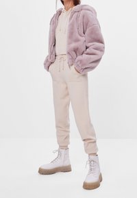 Persoon in een lichtroze fluffy zip-up hoodie, een crèmekleurige hoodie en joggers, met beige chunky veterschoenen, staand tegen een witte achtergrond.