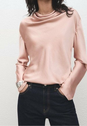 Femme portant un haut à manches longues rose clair avec un col bénitier et un jean bleu foncé taille haute, mains dans les poches, et un bracelet argenté au poignet gauche.