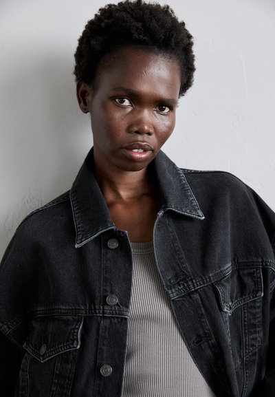 Black denim jacket with a structured silhouette, featuring large pockets and metal snap buttons, layered over a gray ribbed tank top.