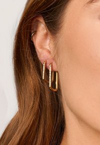 Boucles d'oreilles en ton doré présentant un design géométrique ouvert avec deux barres verticales et de petits cristaux transparents incrustés le long d'une barre.