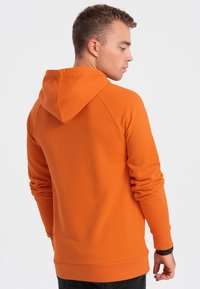 Felpa arancione con una vestibilità rilassata, dotata di cappuccio con cordino e polsini a costine. La texture del tessuto appare liscia ed elastica.