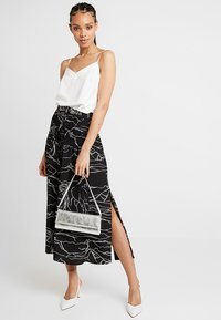 Witte satijnen camisole met dunne bandjes, gecombineerd met een zwarte maxi rok met witte abstracte lijnen en zijsplitten. Zilveren handtas.