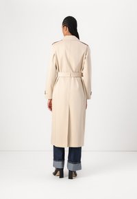 Trench coat bej cu o curea, mâneci lungi și ventilație dorsală. Material neted, guler clasic și epoleți pe umeri. Purtați peste blugi cu manșete.